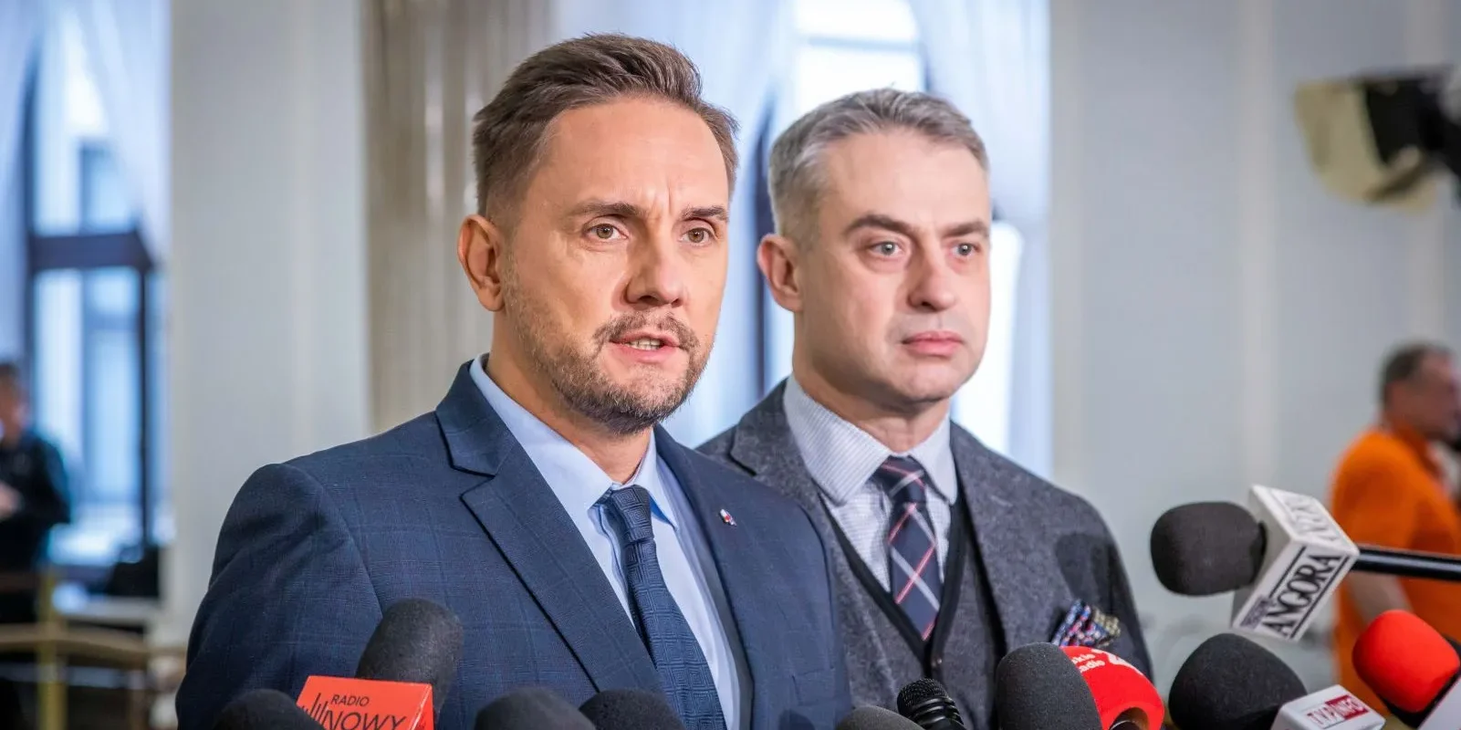 Dwóch mężczyzn w eleganckich strojach stoi przed mikrofonami podczas konferencji prasowej w jasnym pomieszczeniu.