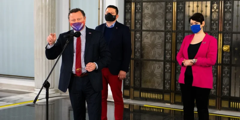 Trzy osoby w maseczkach stoją w eleganckim holu z ozdobnymi drzwiami; mężczyzna po lewej przemawia do mikrofonu, drugi mężczyzna stoi za nim, a kobieta w różowym żakiecie stoi po prawej.