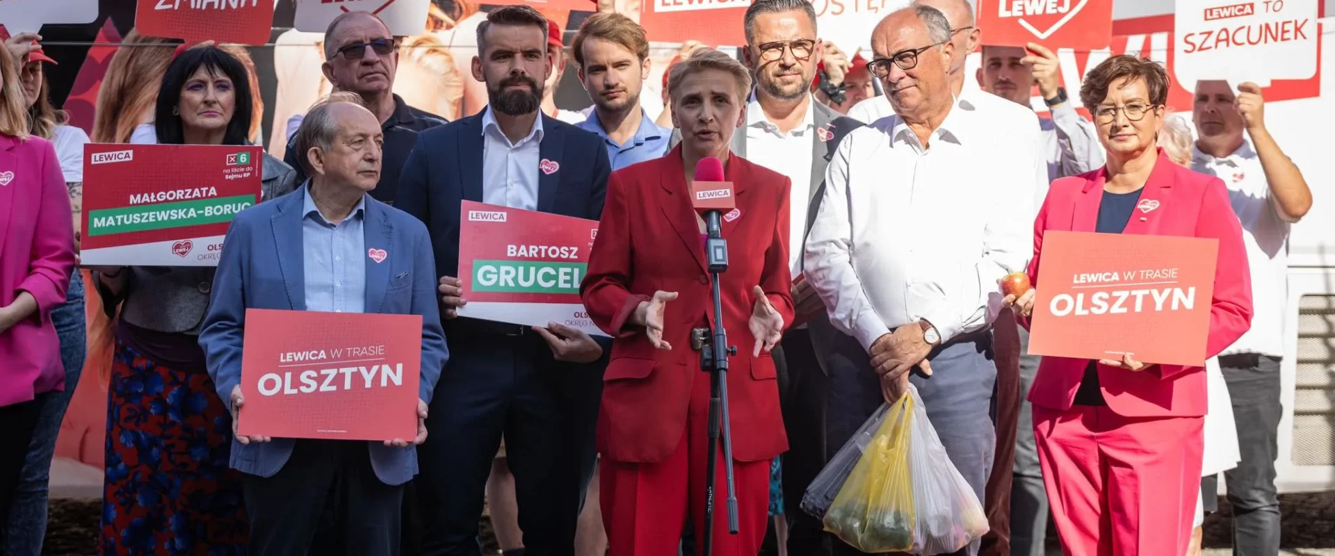 Grupa działaczy Lewicy na wiecu wyborczym, na pierwszym planie kobieta w czerwonym garniturze przemawia do mikrofonu, otoczona innymi politykami trzymającymi plakaty z napisem OLSZTYN.