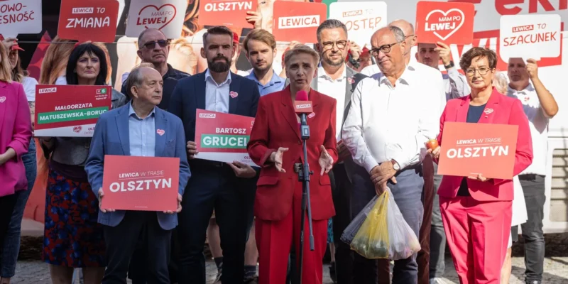 Grupa działaczy Lewicy na wiecu wyborczym, na pierwszym planie kobieta w czerwonym garniturze przemawia do mikrofonu, otoczona innymi politykami trzymającymi plakaty z napisem OLSZTYN.