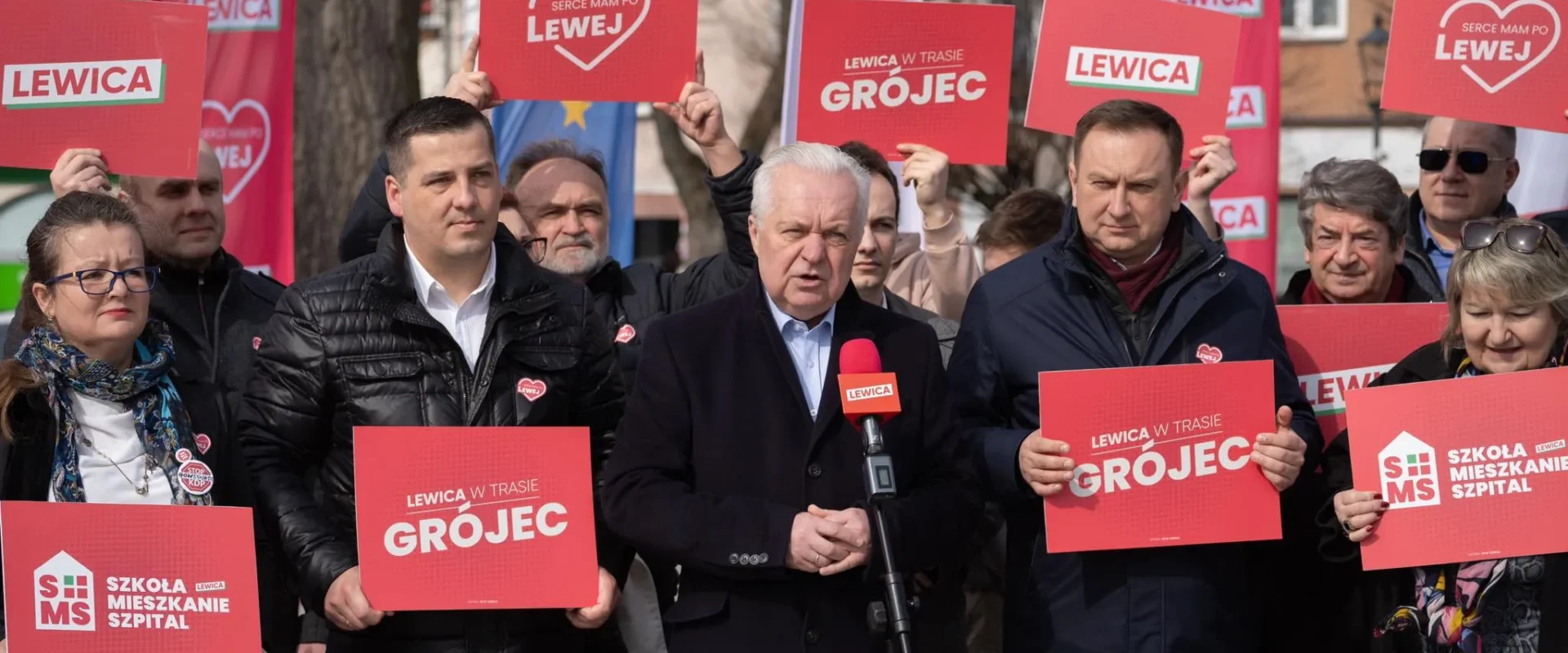 Grupa zwolenników Lewicy na zewnątrz, trzymających czerwone plakaty z napisem LEWICA; w centrum mężczyzna przemawia do mikrofonu podczas wiecu.