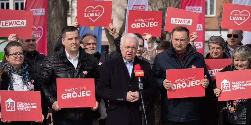 Grupa zwolenników Lewicy na zewnątrz, trzymających czerwone plakaty z napisem LEWICA; w centrum mężczyzna przemawia do mikrofonu podczas wiecu.