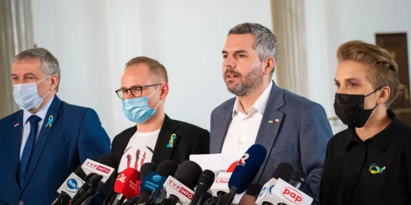 Na zdjęciu widać czwórkę ludzi podczas konferencji prasowej, którzy występują przed mikrofonami w przestrzeni publicznej.