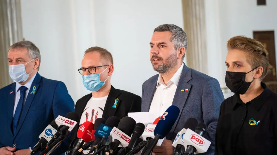 Na zdjęciu widać czwórkę ludzi podczas konferencji prasowej, którzy występują przed mikrofonami w przestrzeni publicznej.