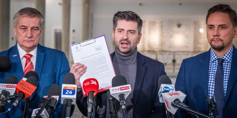 Na zdjęciu trzech mężczyzn stoi przed dziennikarzami, z jednym z nich trzymającym w ręku dokument oraz mówiącym podczas konferencji prasowej.