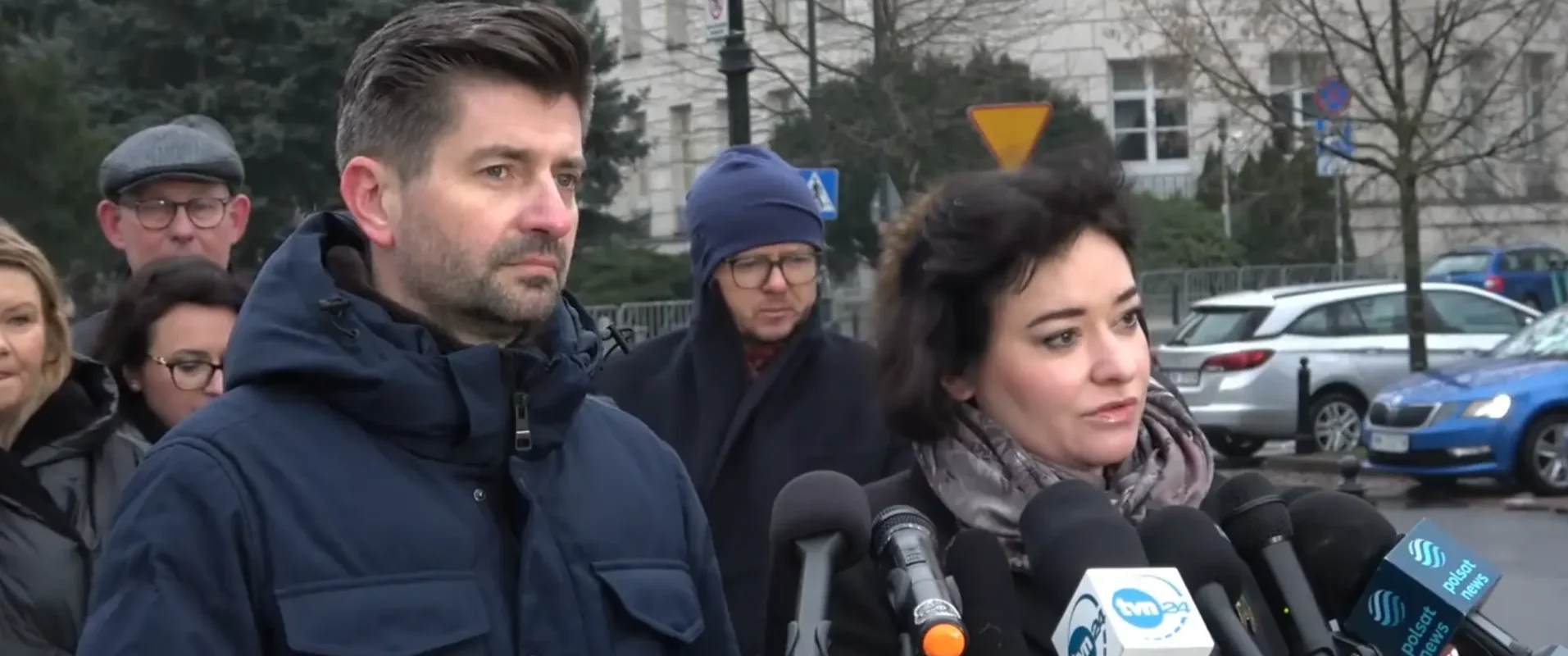 Mężczyzna i kobieta udzielają wywiadu przed mikrofonami podczas konferencji prasowej na zewnątrz, w tle stoją inni uczestnicy i ulica z samochodami.