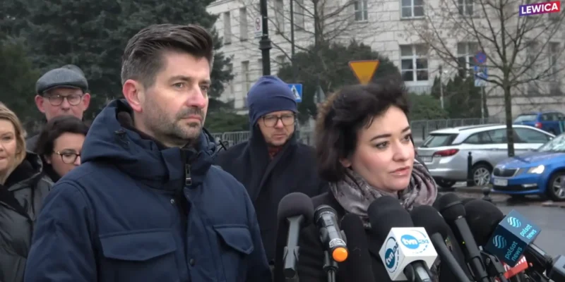 Mężczyzna i kobieta udzielają wywiadu przed mikrofonami podczas konferencji prasowej na zewnątrz, w tle stoją inni uczestnicy i ulica z samochodami.