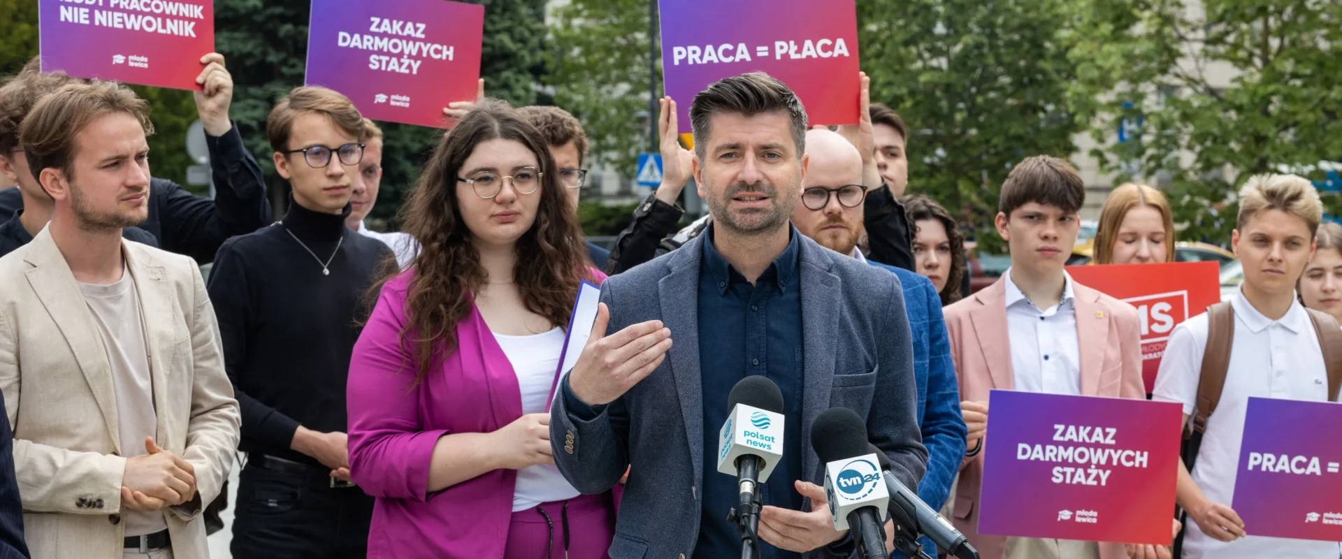 Grupa młodych ludzi trzyma kolorowe plakaty z hasłami „Zakaz darmowych staży” i „Praca = Płaca” podczas konferencji prasowej, centralnie przemawia do mikrofonów mężczyzna.