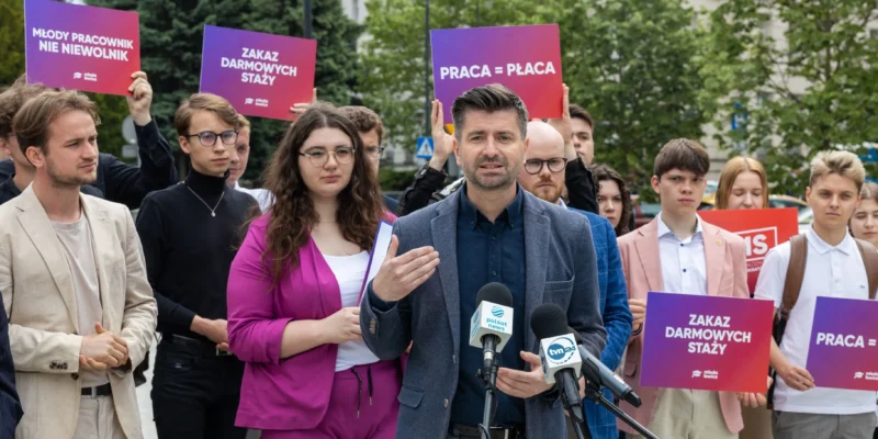 Grupa młodych ludzi trzyma kolorowe plakaty z hasłami „Zakaz darmowych staży” i „Praca = Płaca” podczas konferencji prasowej, centralnie przemawia do mikrofonów mężczyzna.