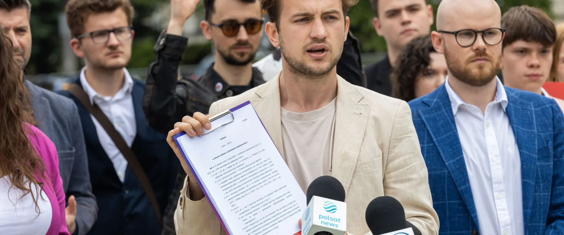 Mężczyzna w beżowym garniturze trzyma clipboard z dokumentami i przemawia do mediów podczas konferencji prasowej, w otoczeniu reporterów i mikrofonów z logotypami Polsat News i TVN24.