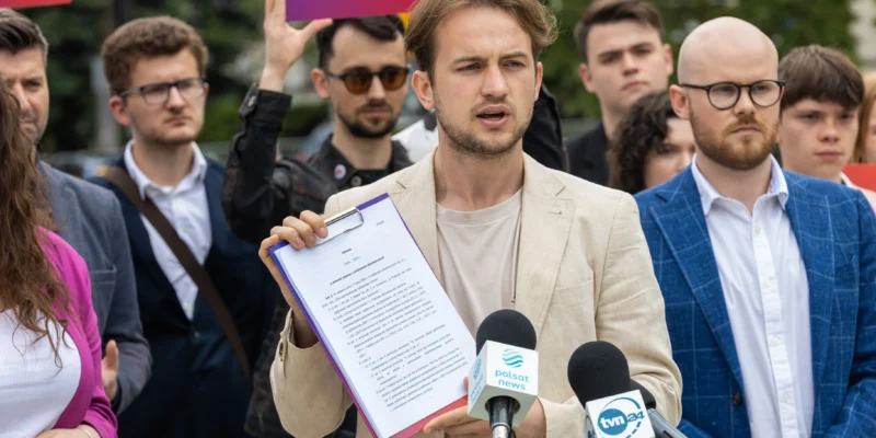 Mężczyzna w beżowym garniturze trzyma clipboard z dokumentami i przemawia do mediów podczas konferencji prasowej, w otoczeniu reporterów i mikrofonów z logotypami Polsat News i TVN24.