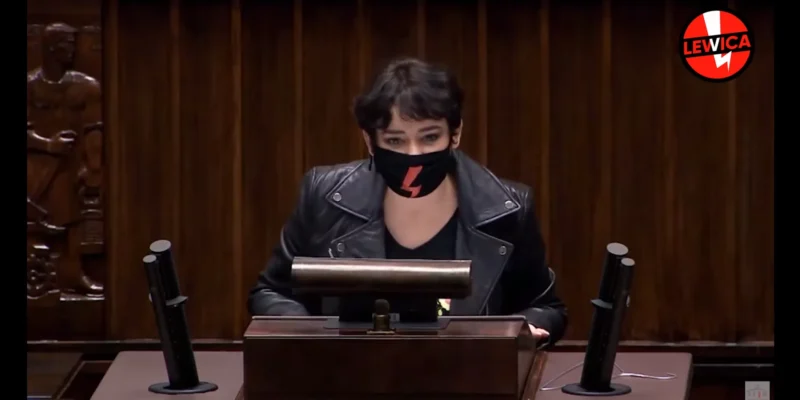 Na zdjęciu widoczna jest kobieta mówiąca za mównicą, ubrana w skórzaną kurtkę i maseczkę z charakterystycznym symbolem, w tle wyraźnie widać drewnianą ścianę z rzeźbami.