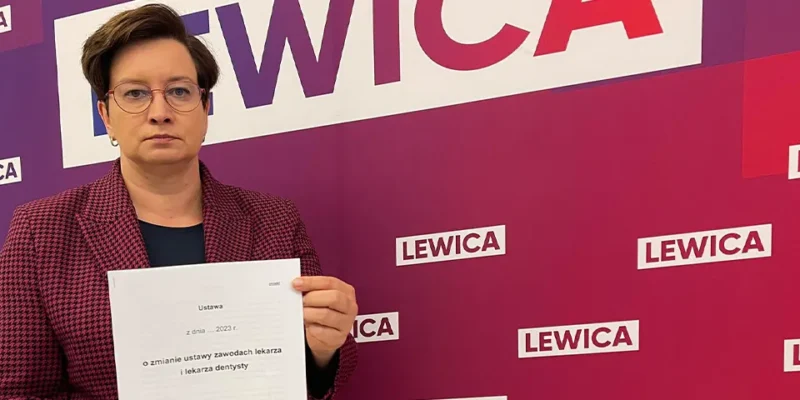 Kobieta w okularach trzyma dokument na tle banerów Lewicy, odnoszących się do projektu uchwały znoszącej klauzulę sumienia.