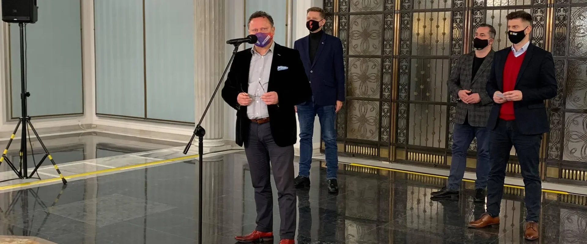 Mężczyzna w garniturze przemawia do mikrofonu w eleganckim holu, za nim stoją trzej mężczyźni w marynarkach, wszyscy w maseczkach, na tle ozdobnych drzwi i marmurowej podłogi.