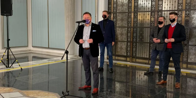 Mężczyzna w garniturze przemawia do mikrofonu w eleganckim holu, za nim stoją trzej mężczyźni w marynarkach, wszyscy w maseczkach, na tle ozdobnych drzwi i marmurowej podłogi.