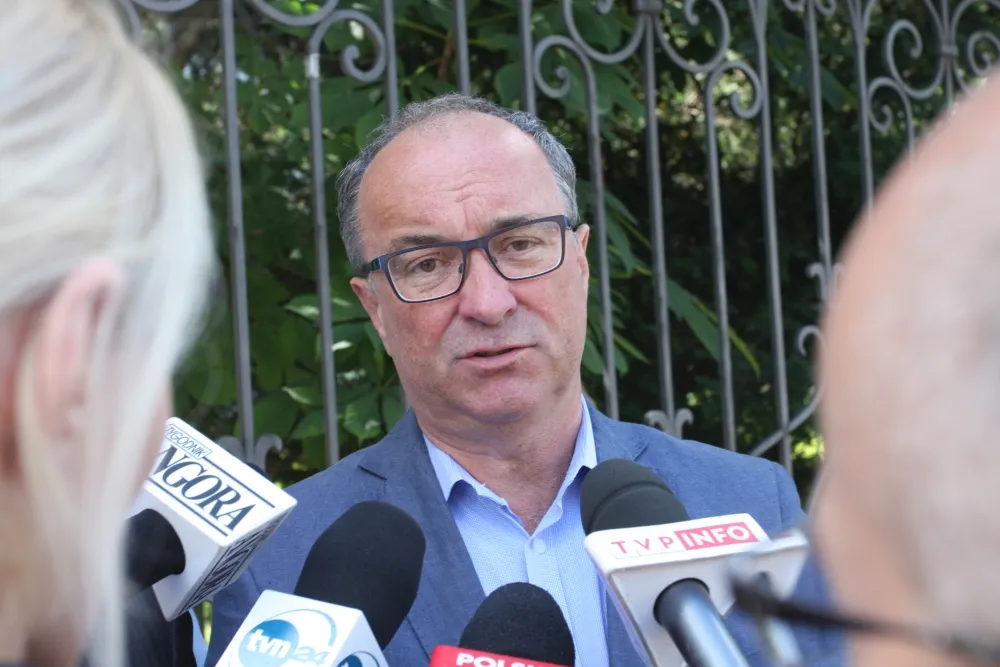 Mężczyzna w okularach, ubrany w niebieską marynarkę, udziela wywiadu przed mikrofonami w otoczeniu zieleni.