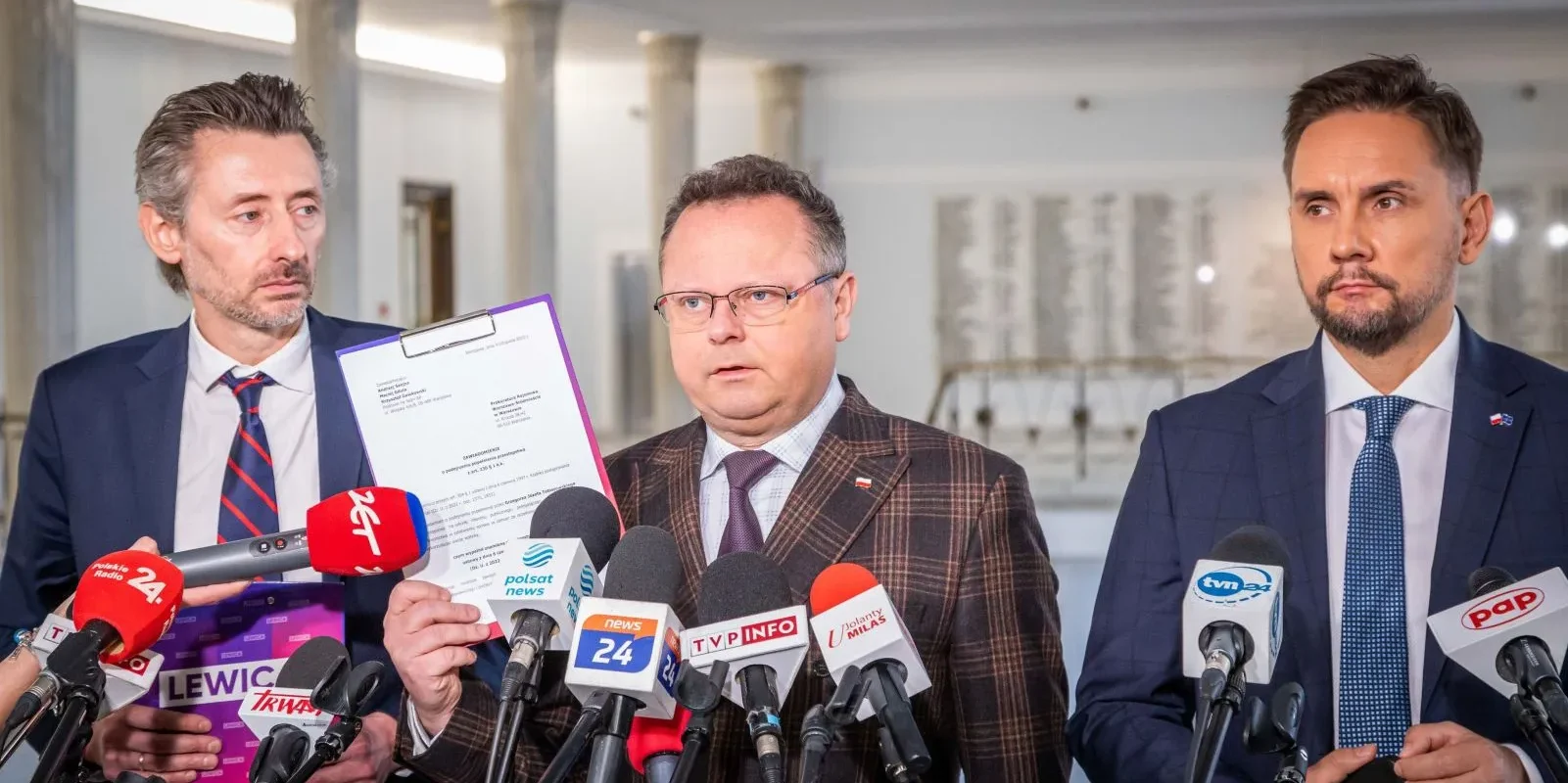 Na zdjęciu trzech mężczyzn stoi przed mikrofonami na konferencji prasowej, z jednym z nich trzymającym dokument.