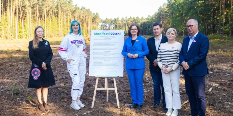 Grupa sześciu osób stoi na leśnej polanie z lasem w tle, obok tablicy z napisem „Manifest 100 dni dla polskiej przyrody i klimatu”.