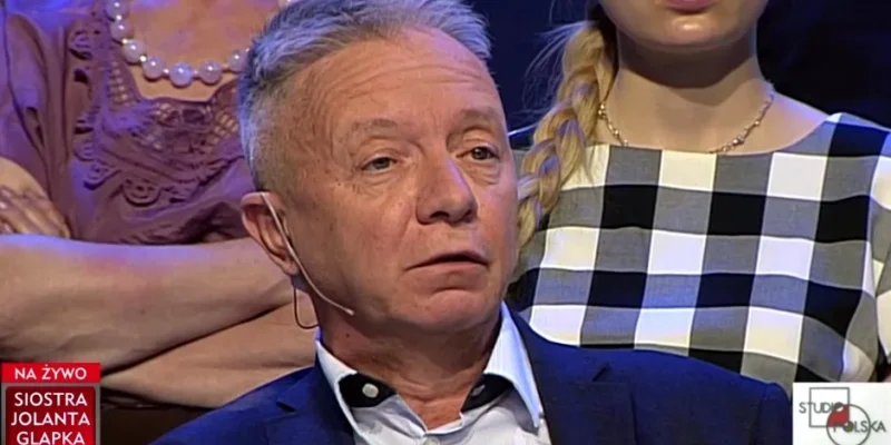 Mężczyzna w garniturze z mikrofonem przy uchu wypowiada się w studiu telewizyjnym przed siedzącą publicznością, z widocznym paskiem informacyjnym u dołu ekranu.