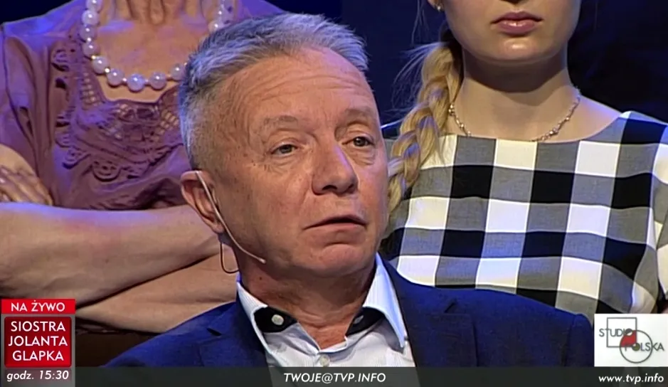 Mężczyzna w garniturze z mikrofonem przy uchu wypowiada się w studiu telewizyjnym przed siedzącą publicznością, z widocznym paskiem informacyjnym u dołu ekranu.