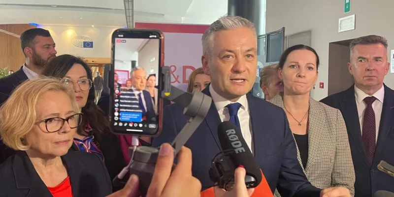 Na zdjęciu widoczna jest grupa osób biorących udział w konferencji prasowej, z prominentnym mówcą na pierwszym planie, otoczonym przez innych uczestników i dziennikarzy.