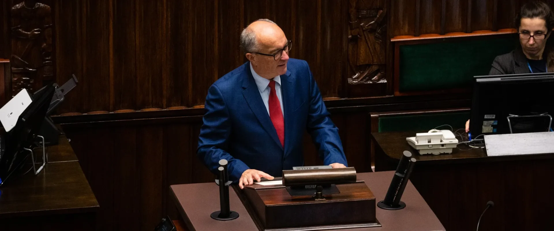 Polityk w niebieskim garniturze i czerwonym krawacie stoi przy mównicy w sali plenarnej, w otoczeniu drewnianych paneli i sprzętu biurowego.