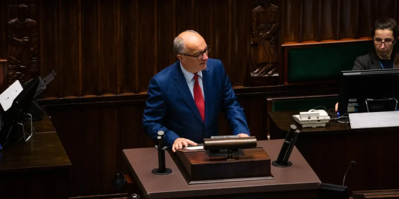 Polityk w niebieskim garniturze i czerwonym krawacie stoi przy mównicy w sali plenarnej, w otoczeniu drewnianych paneli i sprzętu biurowego.