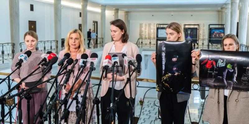 Grupa kobiet stoi na konferencji prasowej w wnętrzu z kolumnami, przed licznymi mikrofonami trzyma plakaty ze zdjęciami uchodźców i apeluje o przywrócenie ładu prawnego na granicy polsko-białoruskiej.