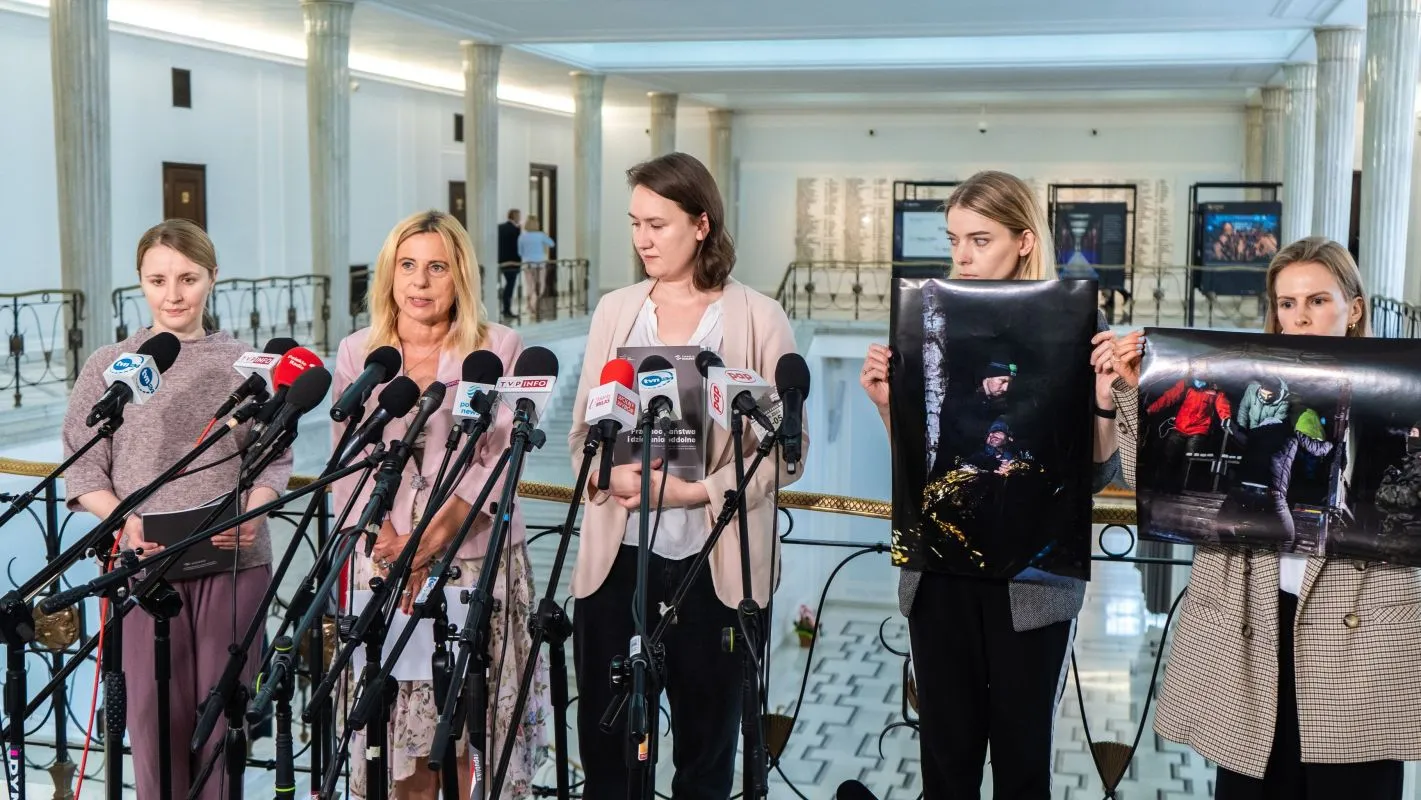 Grupa kobiet stoi na konferencji prasowej w wnętrzu z kolumnami, przed licznymi mikrofonami trzyma plakaty ze zdjęciami uchodźców i apeluje o przywrócenie ładu prawnego na granicy polsko-białoruskiej.