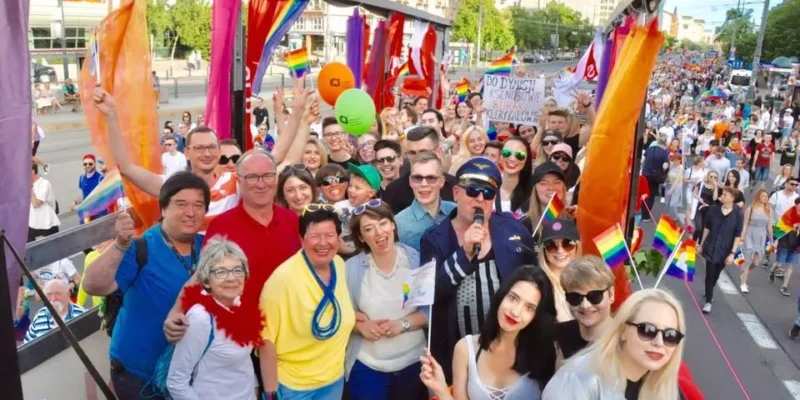 Grupa uczestników SLD pozująca na udekorowanej tęczowymi flagami platformie podczas Parady Równości, z licznym tłumem maszerujących w tle.