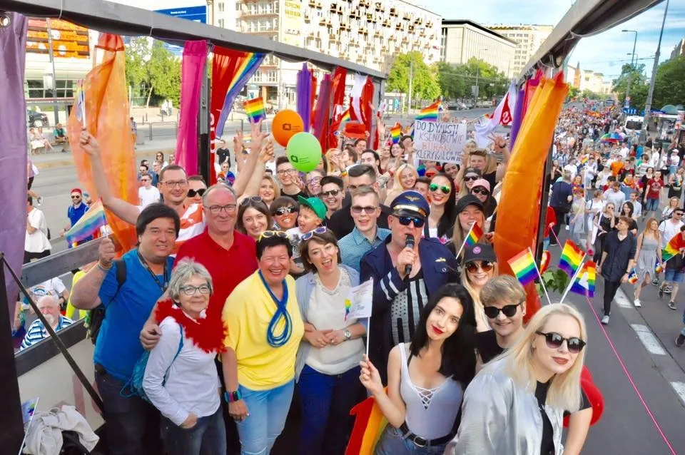Grupa uczestników SLD pozująca na udekorowanej tęczowymi flagami platformie podczas Parady Równości, z licznym tłumem maszerujących w tle.