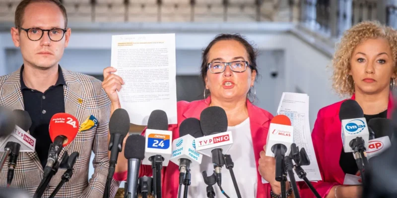 Na zdjęciu widoczna jest trójka ludzi podczas konferencji prasowej, z jedną osobą pokazującą dokument, a przed nimi stoi wiele mikrofonów mediów.
