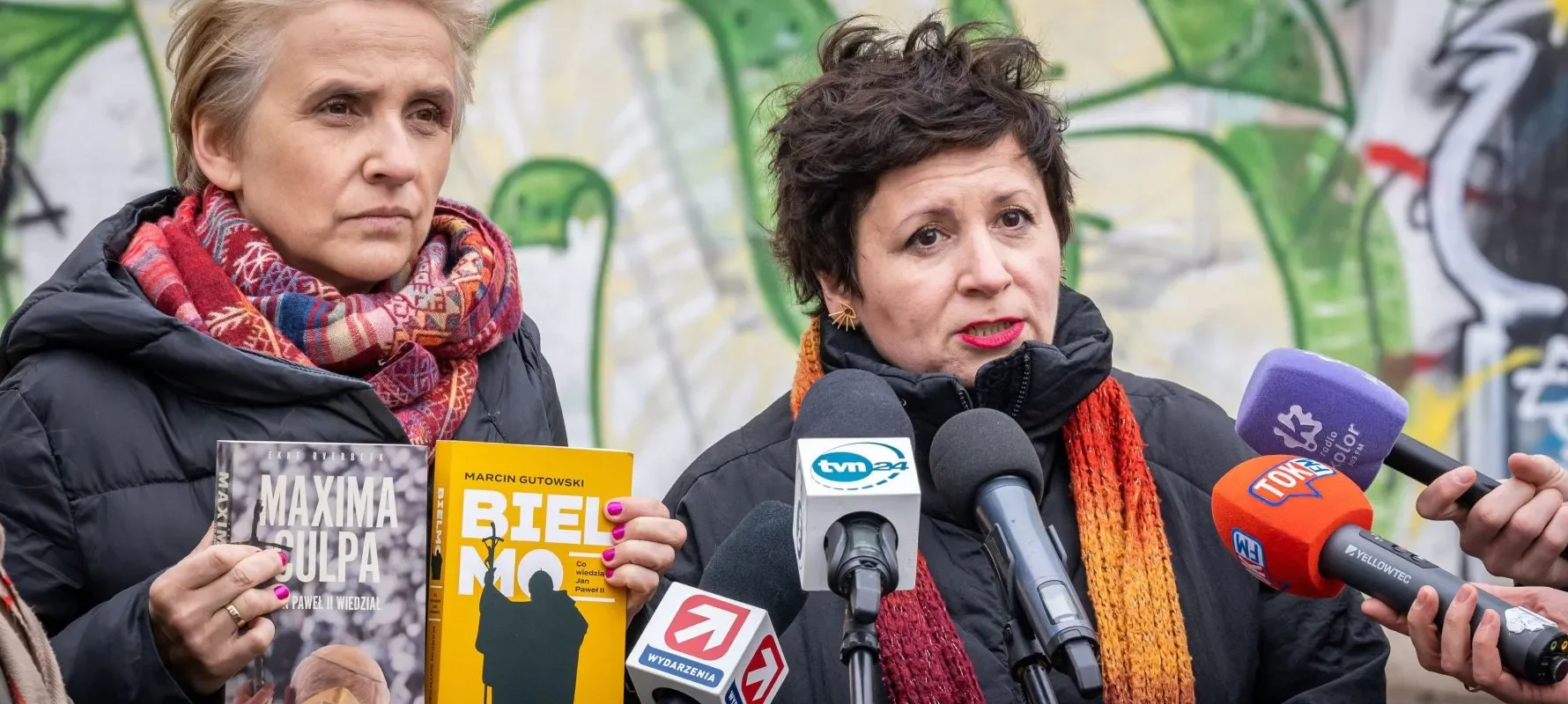 Na zdjęciu dwie kobiety, jedna trzymająca książkę "Maxima Culpa", a druga "Biel Mo", przemawiają publicznie w otoczeniu mediów, na tle kolorowej graffiti.
