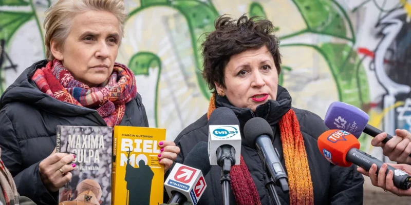 Na zdjęciu dwie kobiety, jedna trzymająca książkę "Maxima Culpa", a druga "Biel Mo", przemawiają publicznie w otoczeniu mediów, na tle kolorowej graffiti.