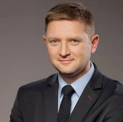 Portret mężczyzny w garniturze i krawacie, lekko uśmiechniętego, na neutralnym szarym tle.