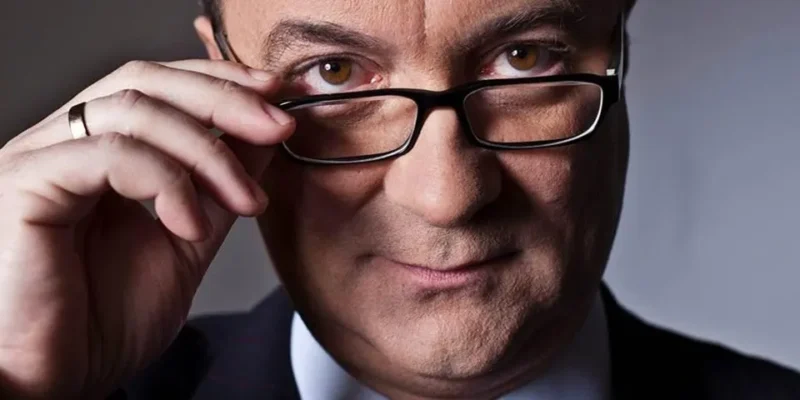 Portret mężczyzny w okularach, z lekkim uśmiechem, trzymającego okulary dłonią.