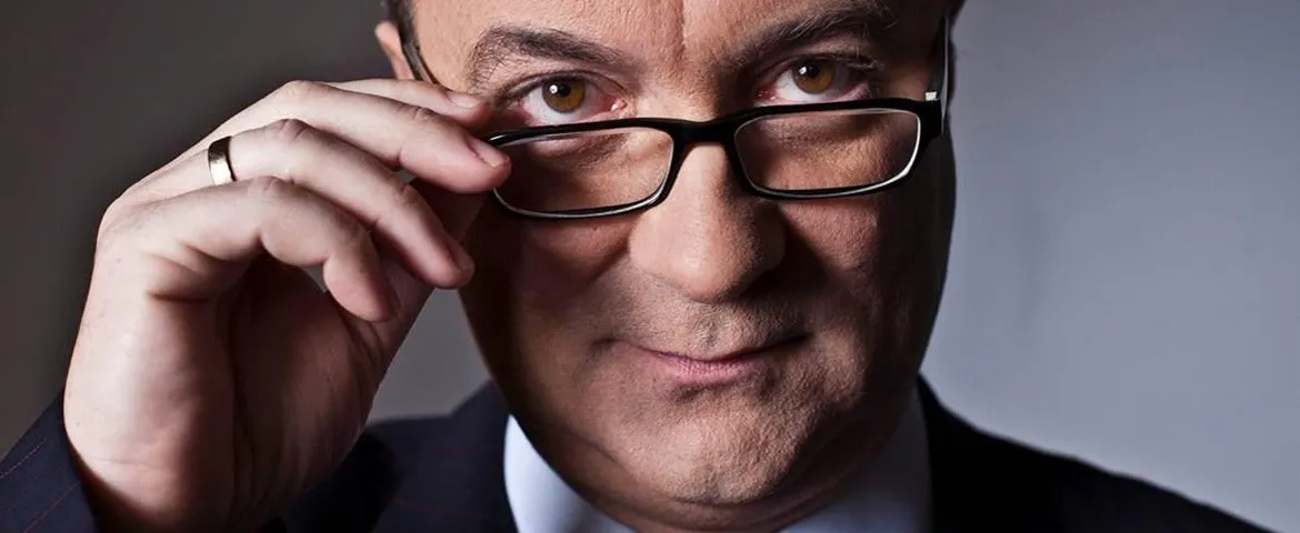 Portret mężczyzny w okularach, z lekkim uśmiechem, trzymającego okulary dłonią.