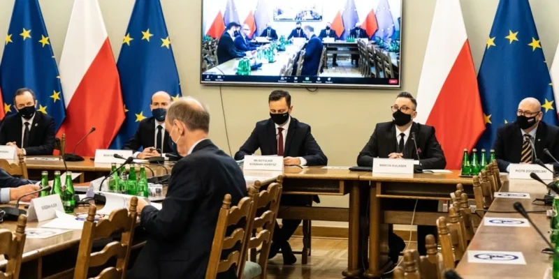 Uczestnicy formalnego spotkania w sali konferencyjnej, wszyscy w maseczkach, siedzą przy długim stole z identyfikatorami, a na tle widać flagi Unii Europejskiej i Polski oraz monitor z wideokonferencją.