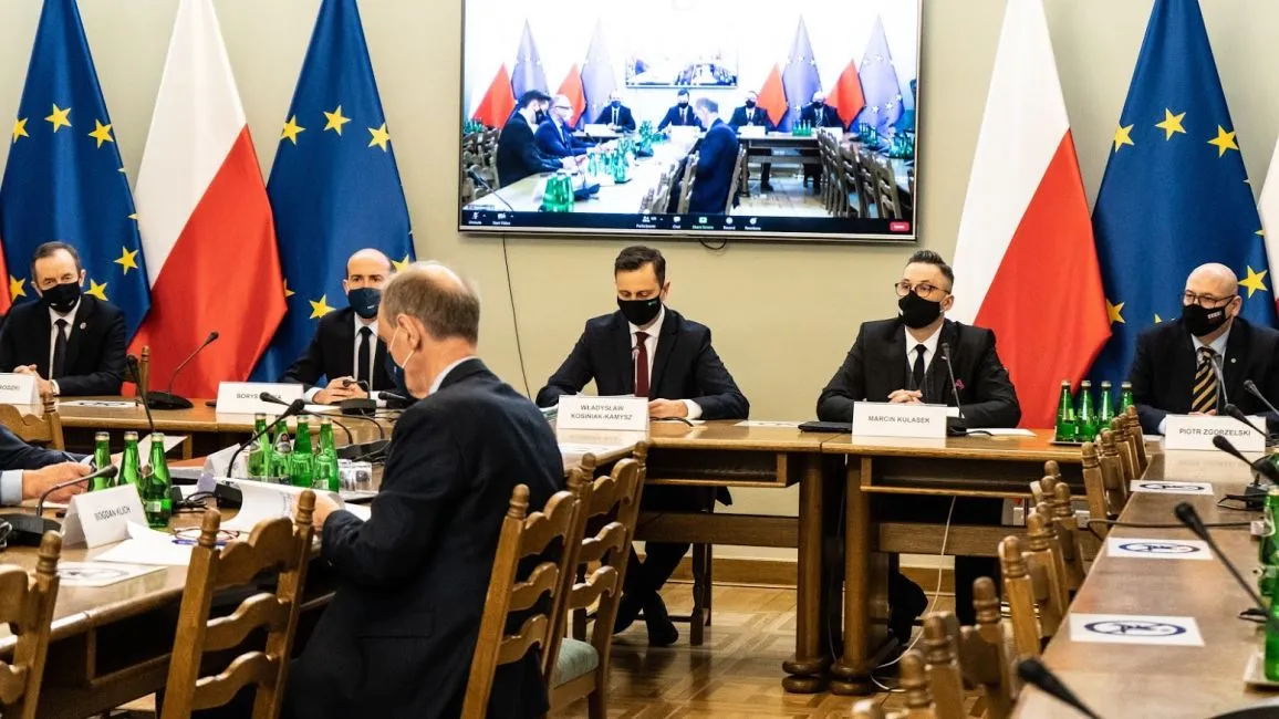 Uczestnicy formalnego spotkania w sali konferencyjnej, wszyscy w maseczkach, siedzą przy długim stole z identyfikatorami, a na tle widać flagi Unii Europejskiej i Polski oraz monitor z wideokonferencją.