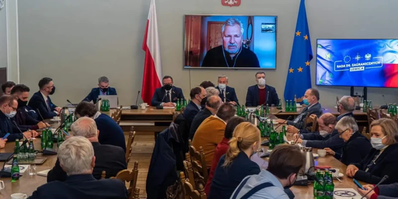 Na zdjęciu przedstawiona jest sala konferencyjna, w której odbywa się spotkanie, a w tle widoczny jest ekran z osobą uczestniczącą w rozmowie online.