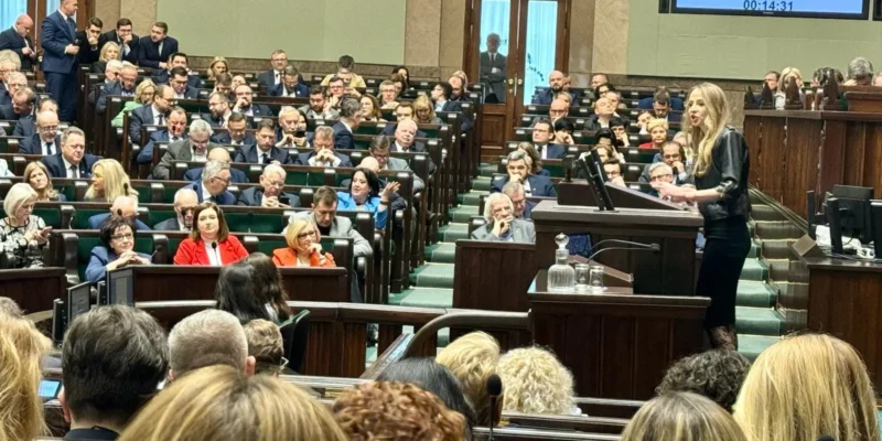 Kobieta wygłasza przemówienie z mównicy w sejmowej sali plenarnej, podczas gdy zgromadzeni posłowie siedzą i słuchają.