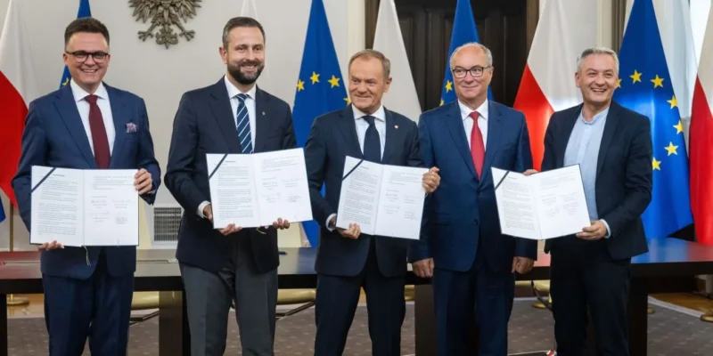 Pięciu mężczyzn w garniturach trzyma podpisane dokumenty, stojąc przed flagami Polski i Unii Europejskiej podczas wydarzenia koalicji szerokiego kompromisu.
