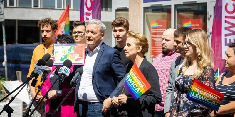 Grupa osób podczas konferencji prasowej na Paradzie Równości 2022 stoi przy mikrofonach, prezentuje mapę Europy i trzyma tęczowe flagi Lewica.