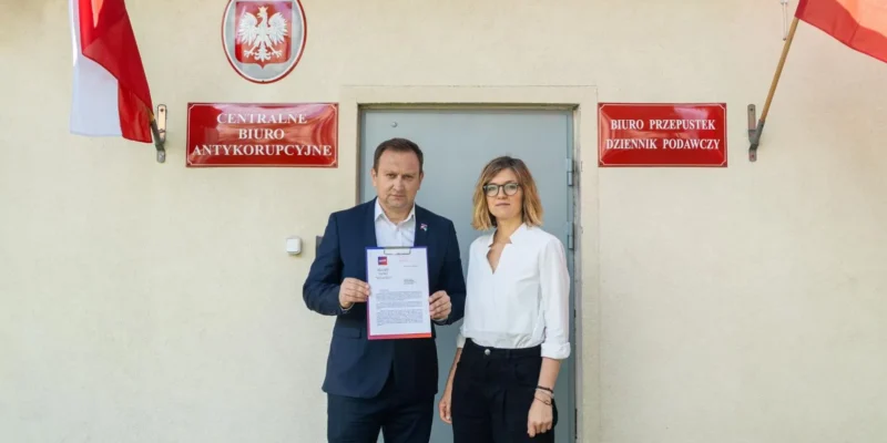 Dwóch ludzi stoi przed wejściem do siedziby Centralnego Biura Antykorupcyjnego, trzymając dokumenty, w otoczeniu flag państwowych.