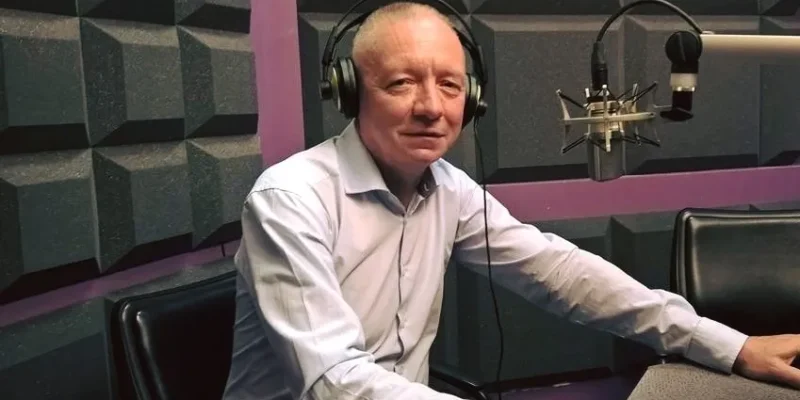 Mężczyzna w koszuli i słuchawkach siedzi przy mikrofonie w studiu radiowym.