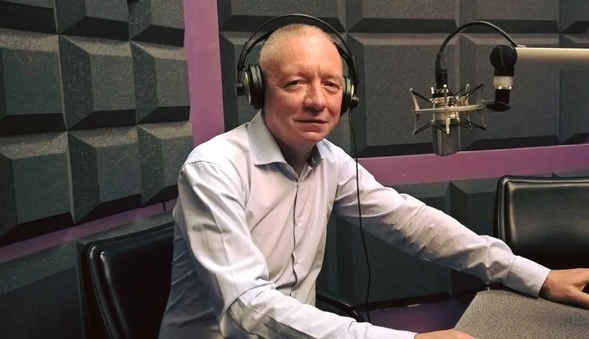 Mężczyzna w koszuli i słuchawkach siedzi przy mikrofonie w studiu radiowym.