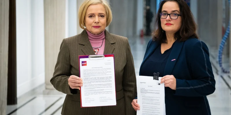 Na zdjęciu dwie kobiety stoją w korytarzu, trzymając dokumenty, z wyraźnym logo Lewicy na jednej z nich.