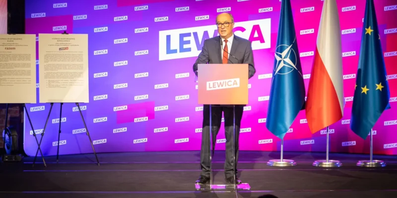 Na zdjęciu widać mężczyznę przemawiającego z podium podczas wydarzenia z logotypem partii Lewica, z flags NATO, Polski i Unii Europejskiej w tle.