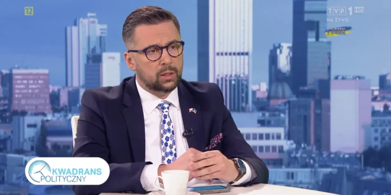 Mężczyzna w eleganckim garniturze siedzi przy stole w programie telewizyjnym, w tle widoczne nowoczesne miasto.