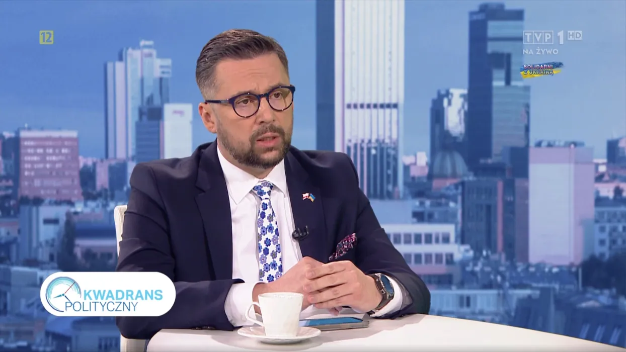 Mężczyzna w eleganckim garniturze siedzi przy stole w programie telewizyjnym, w tle widoczne nowoczesne miasto.